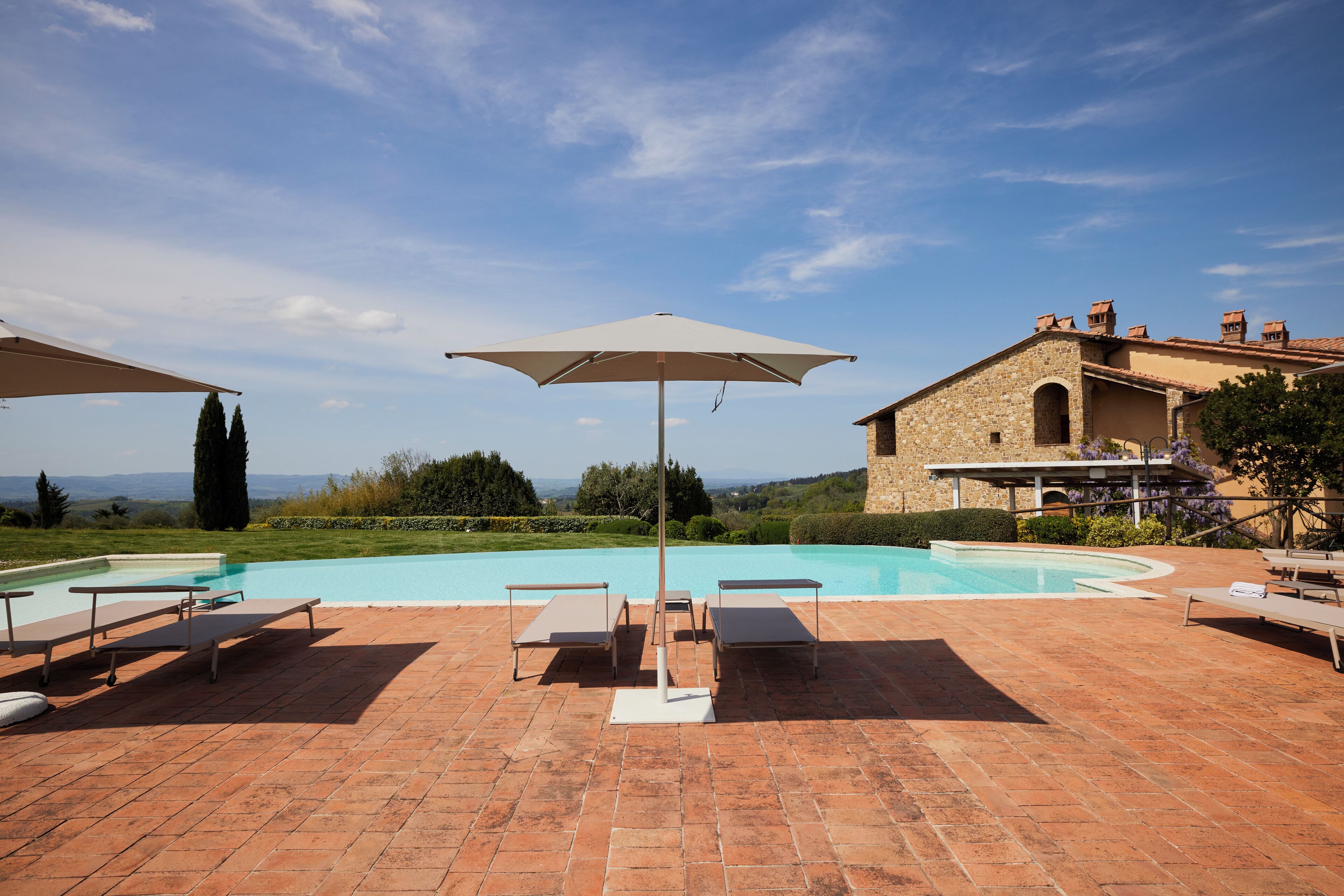 Un espace aménagé au bord de la piscine, avec des chaises longues et un parasol, un bâtiment en pierre et une vue sur le paysage.