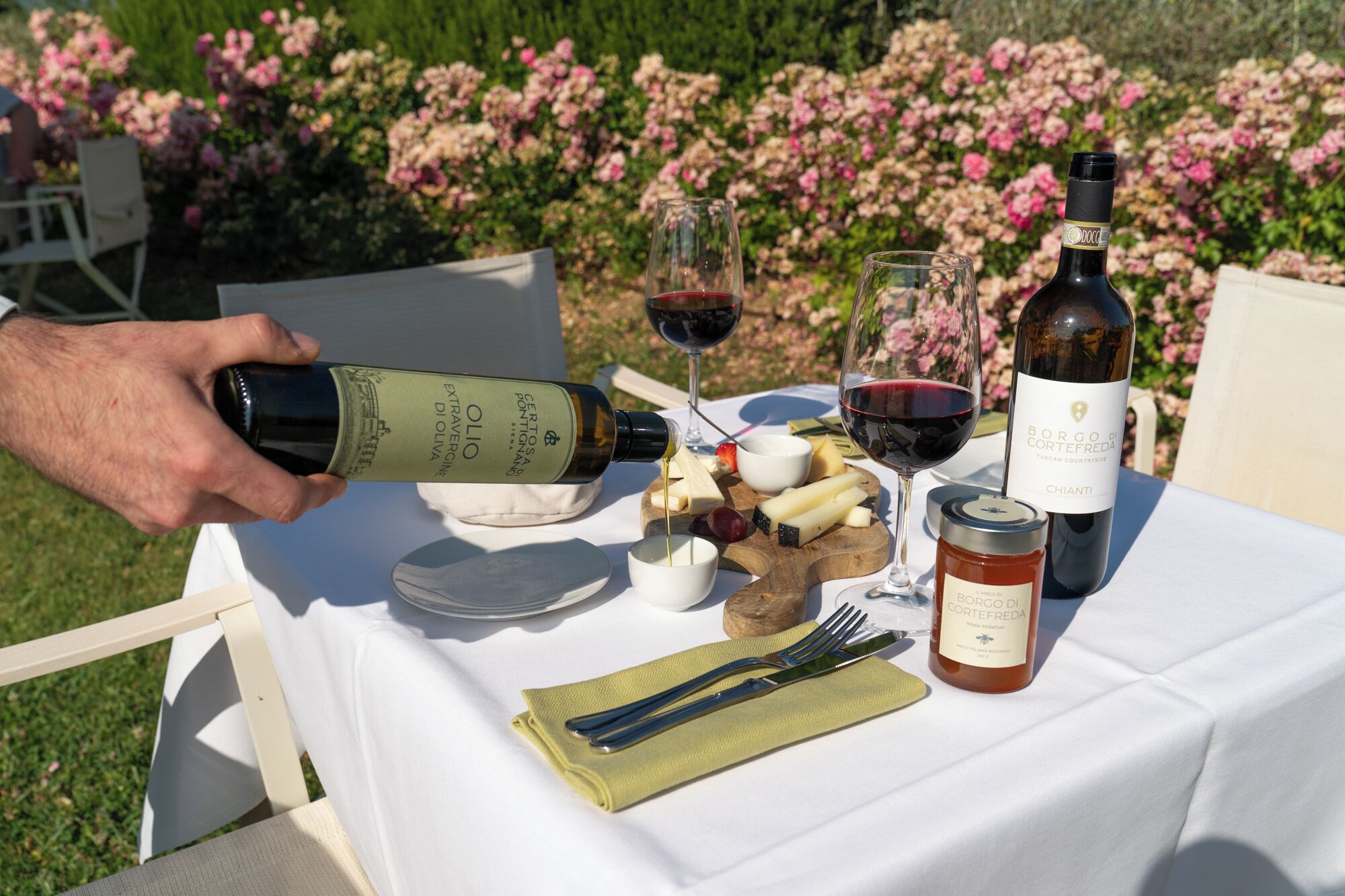 Une table dressée à l’extérieur, avec une bouteille de vin, des verres, du fromage et un pot de confiture.