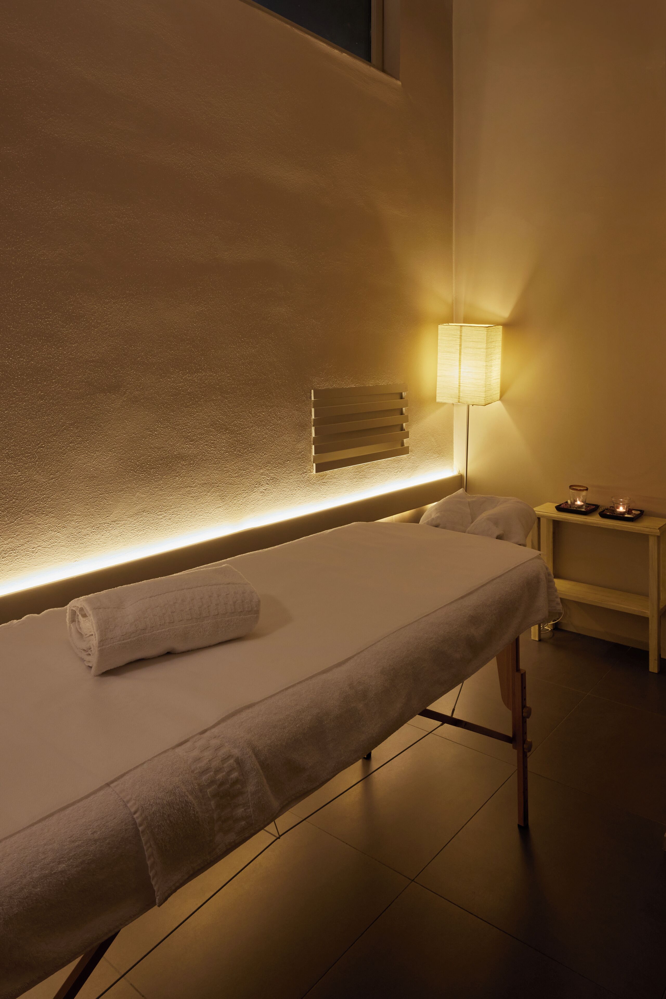 A neatly made bed with a rolled towel, a bedside lamp, and a small table with candles.