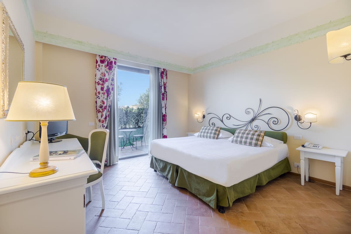 A hotel room with a bed, a desk with a lamp, a chair, a small table, and a telephone.