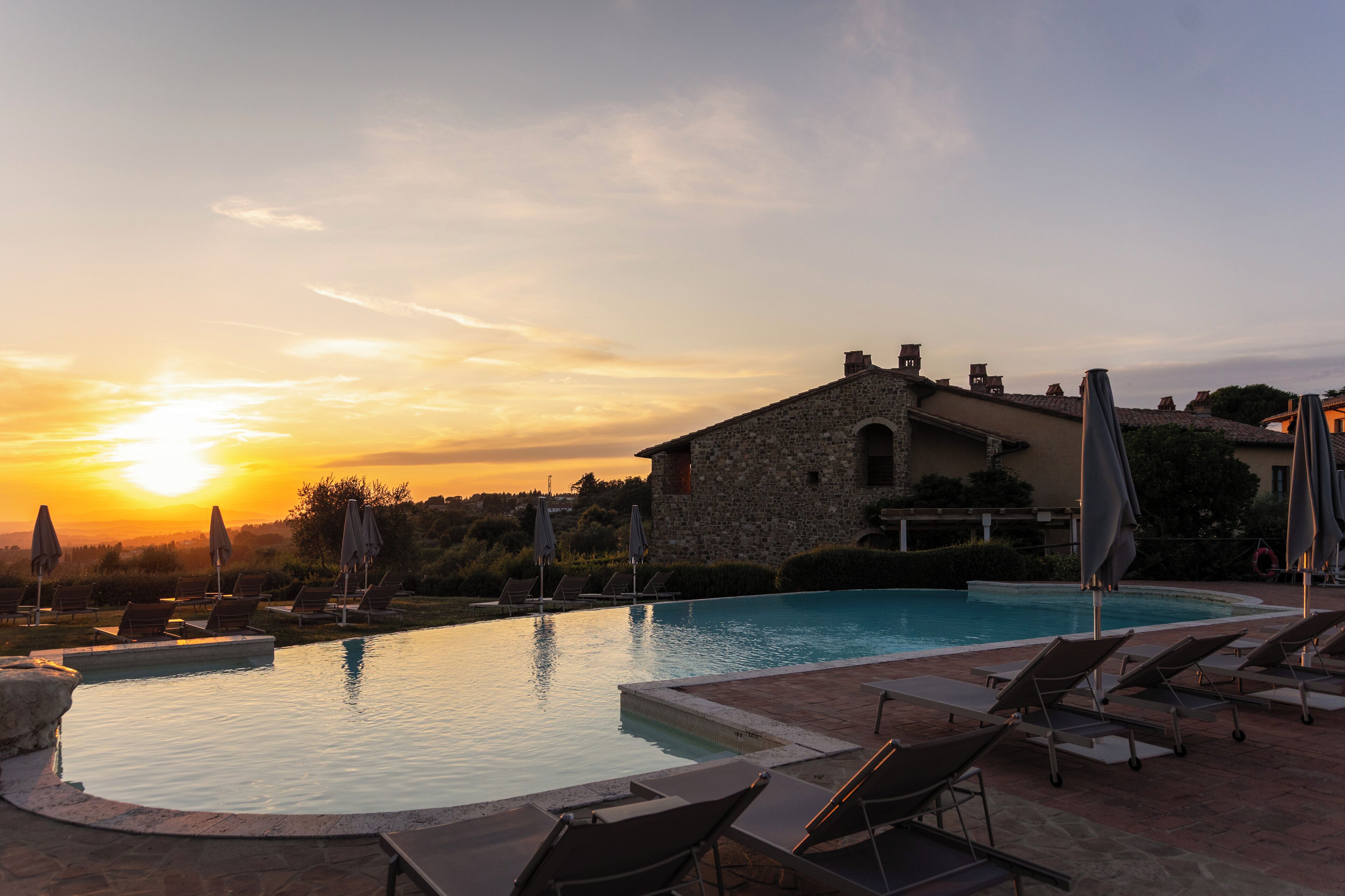 A swimming pool with lounge chairs and umbrellas, a stone building, and a sunset in the background.