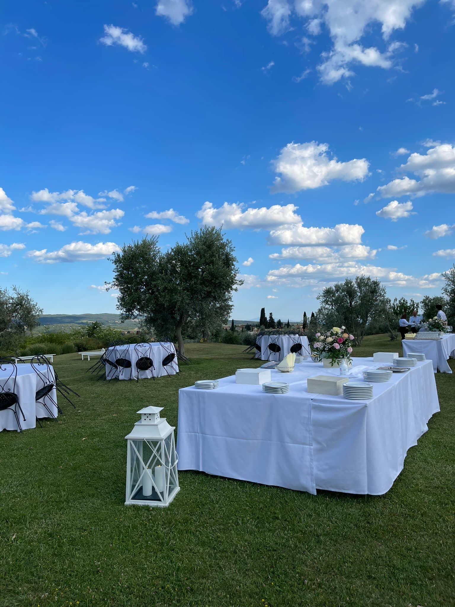 An outdoor event setup with tables covered in white tablecloths, arranged on a grassy field with a clear blue sky and scattered clouds in the background.