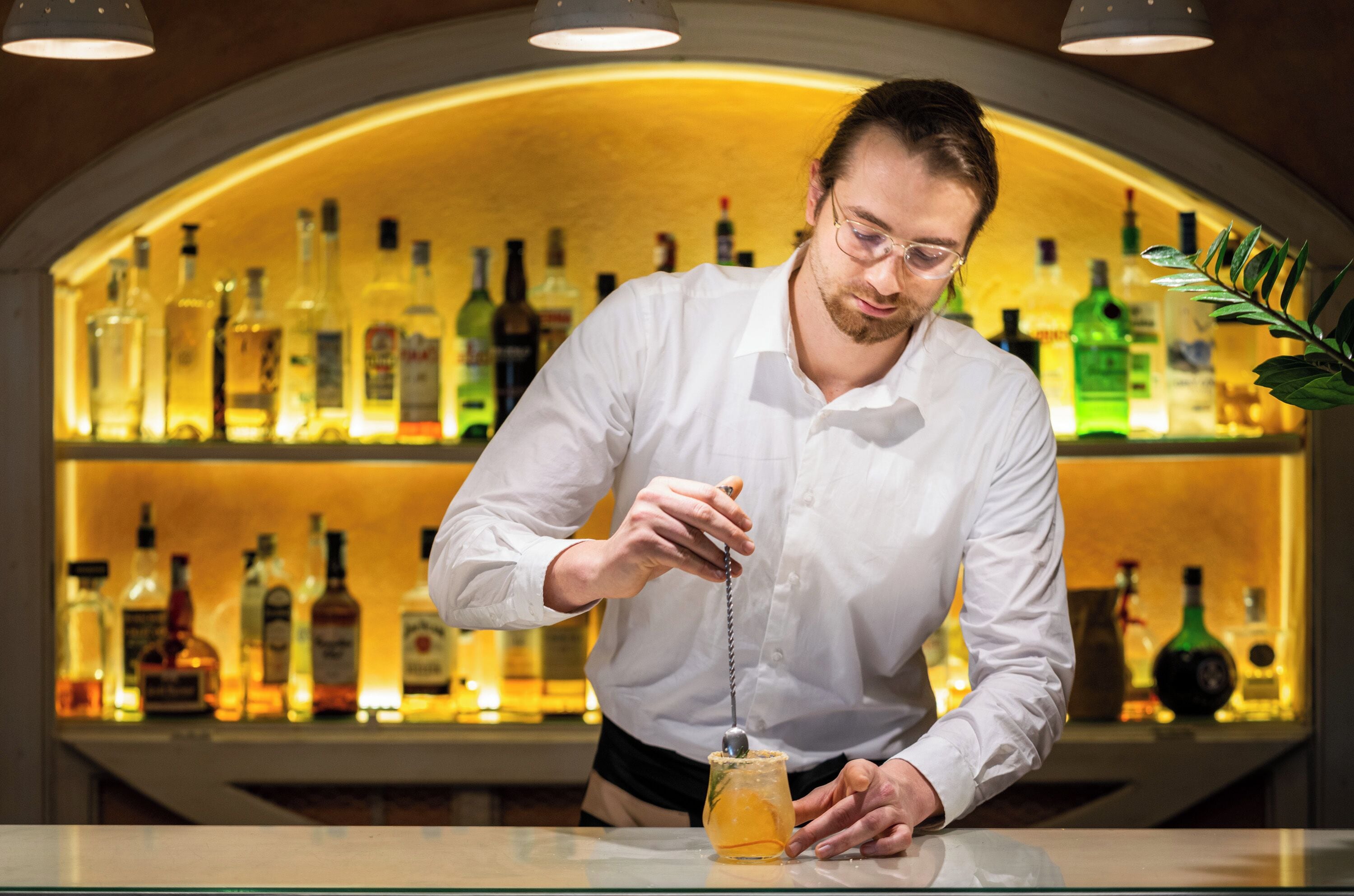 Un barman prépare un cocktail à l’aide d’une cuillère dans un bar, avec des étagères remplies de bouteilles d’alcool en arrière-plan.