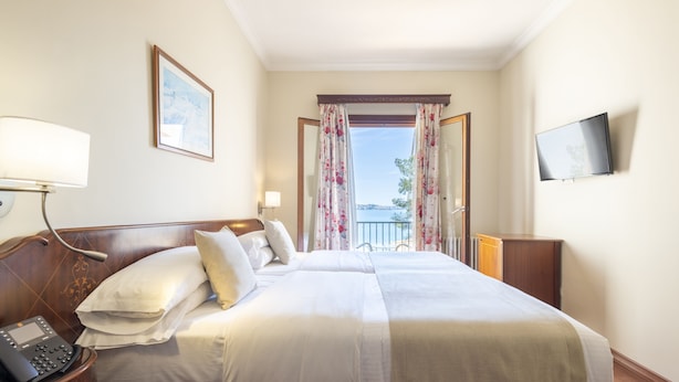 Habitación de hotel con una cama grande, un teléfono en la mesita de noche, un televisor en la pared y vistas al exterior a través de una ventana con cortinas florales.