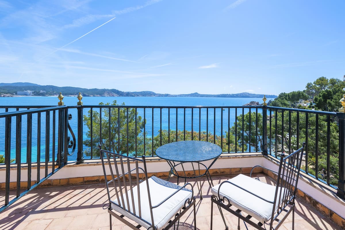A balcony with a table and two chairs, overlooking a body of water and distant land.