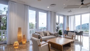 Amplia sala de estar con un sofá grande, dos sillones, una mesa de centro de madera y una mesa de comedor. Ventanas grandes ofrecen vistas a palmeras y un paisaje distante.