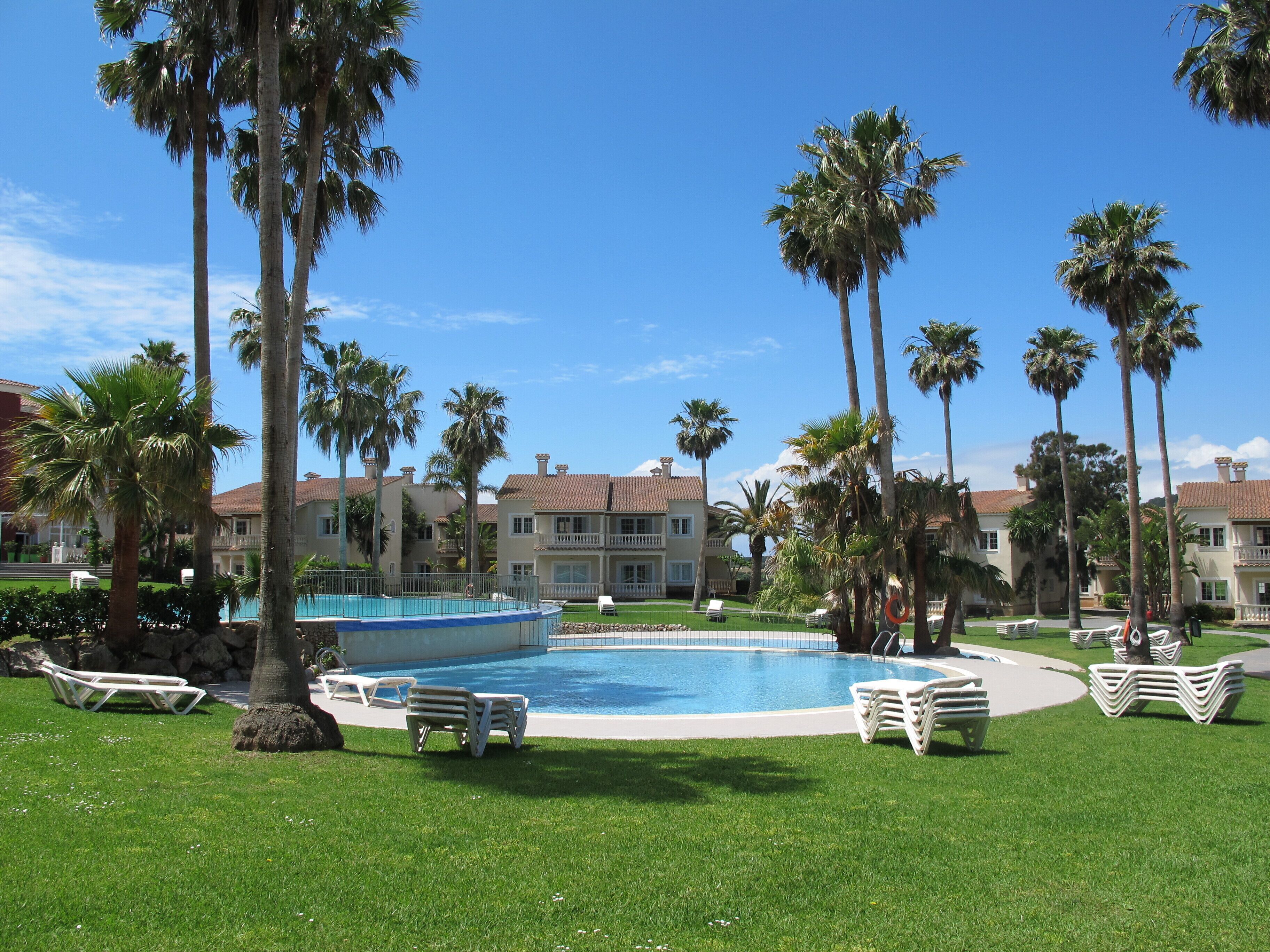Un área de piscina con palmeras, una piscina y un edificio con techo de tejas.