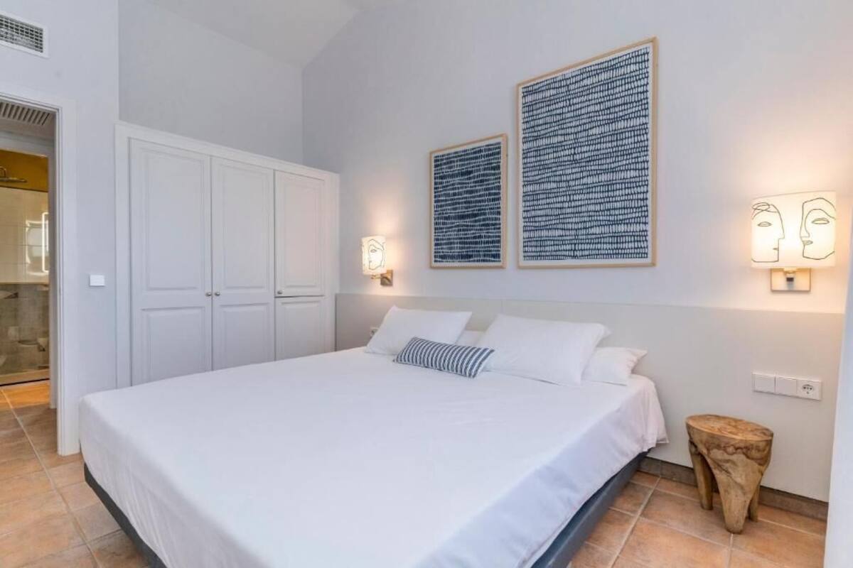 A bedroom with a large bed, white walls, two framed artworks, a wooden stool, and a wardrobe.