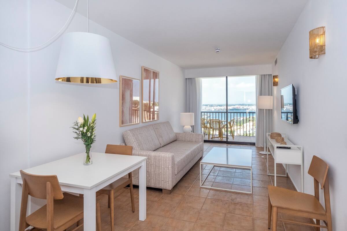 A modern living room with a sofa, dining table, and chairs, a TV, and a balcony with a view.