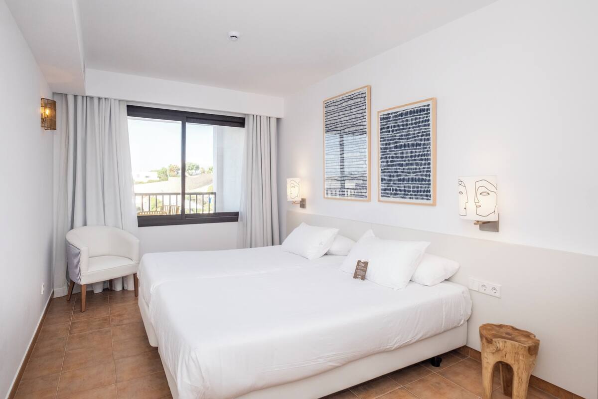 A modern hotel room with a large bed, a chair, a small wooden stool, and two framed artworks on the wall.