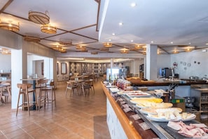 A spacious cafe with multiple seating areas, a buffet counter with various food items, and a modern interior design. - Calallonga Hotel Menorca (Mahón)