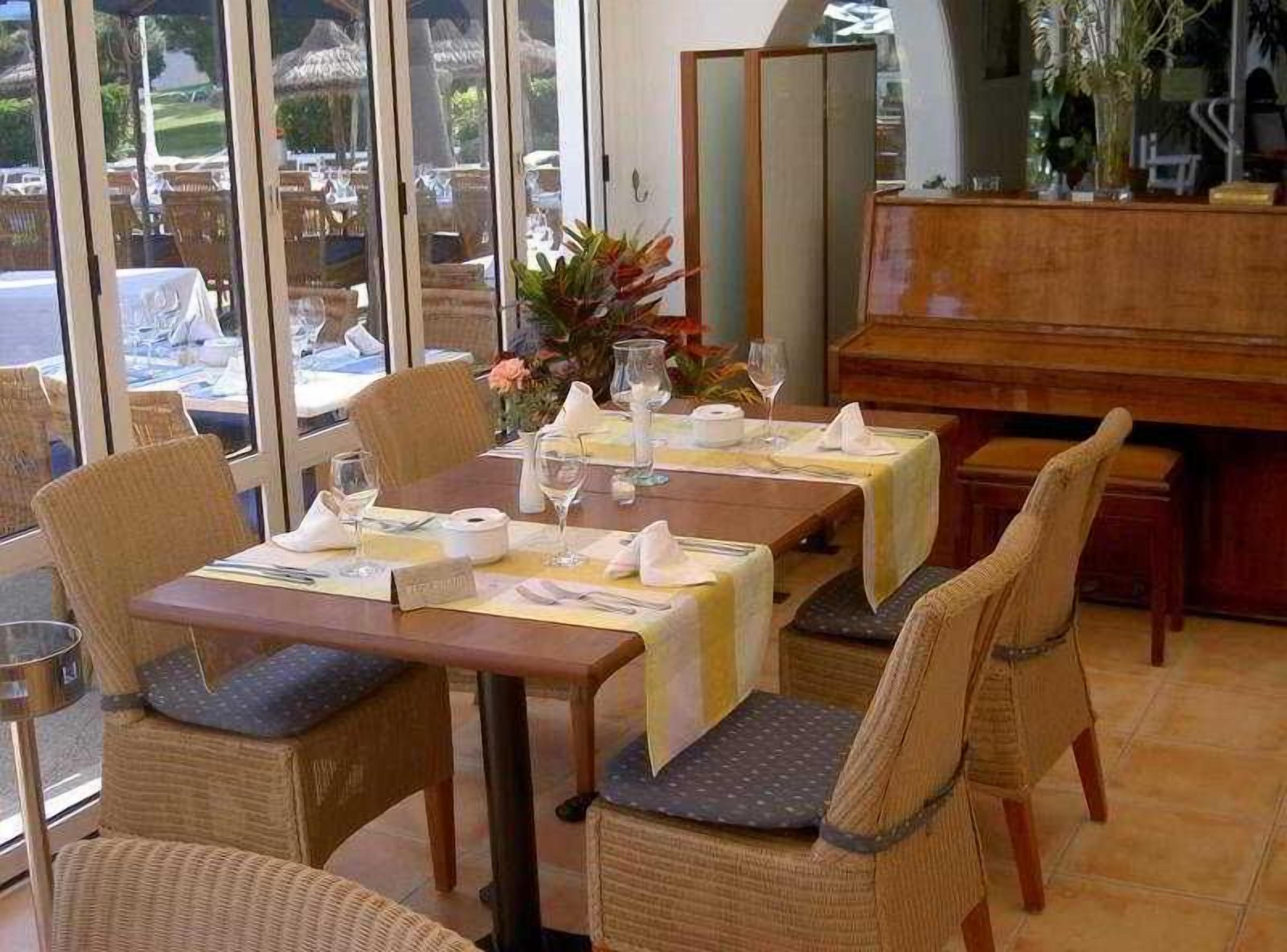 Un comedor con un piano, sillas de mimbre y una mesa para dos, decorada con un jarrón de flores.
