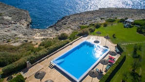 An aerial view of a rectangular swimming pool with a clear blue water surface, surrounded by a paved deck with lounge chairs and umbrellas. - Apartamentos Parque Mar (Santanyi)