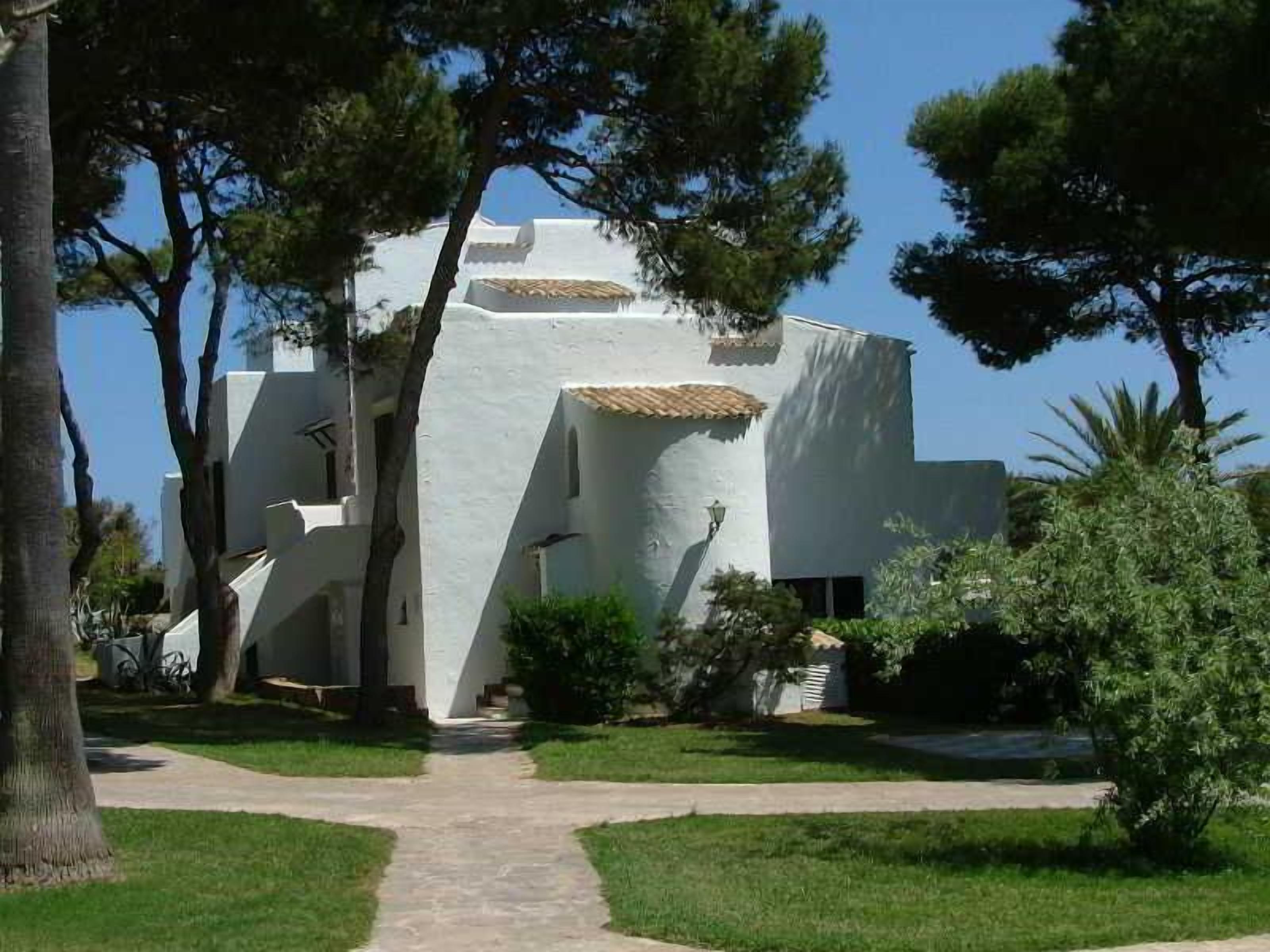 A white building with a pathway leading to it, surrounded by trees and greenery.