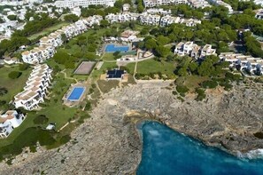 An aerial view of a coastal residential area with multiple white buildings, green spaces, and a rocky shoreline. - Apartamentos Parque Mar (Santanyi)
