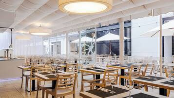 Un'ampia sala da pranzo con tavoli e sedie in legno, grandi lampadari pendenti e vista all'esterno grazie a ampie finestre.