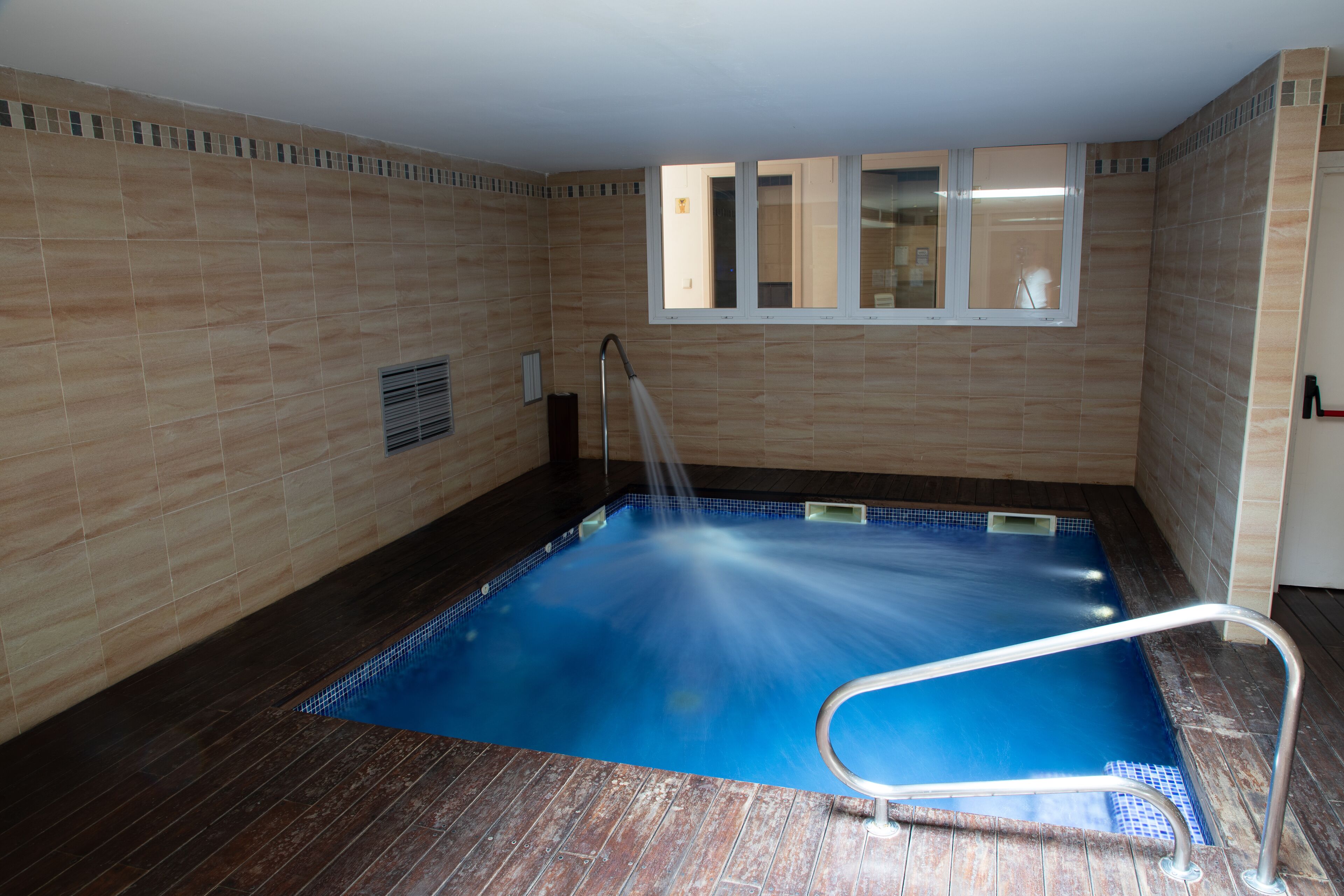 Piscina interior pequena com escada de metal, chão de madeira e paredes revestidas de azulejos.