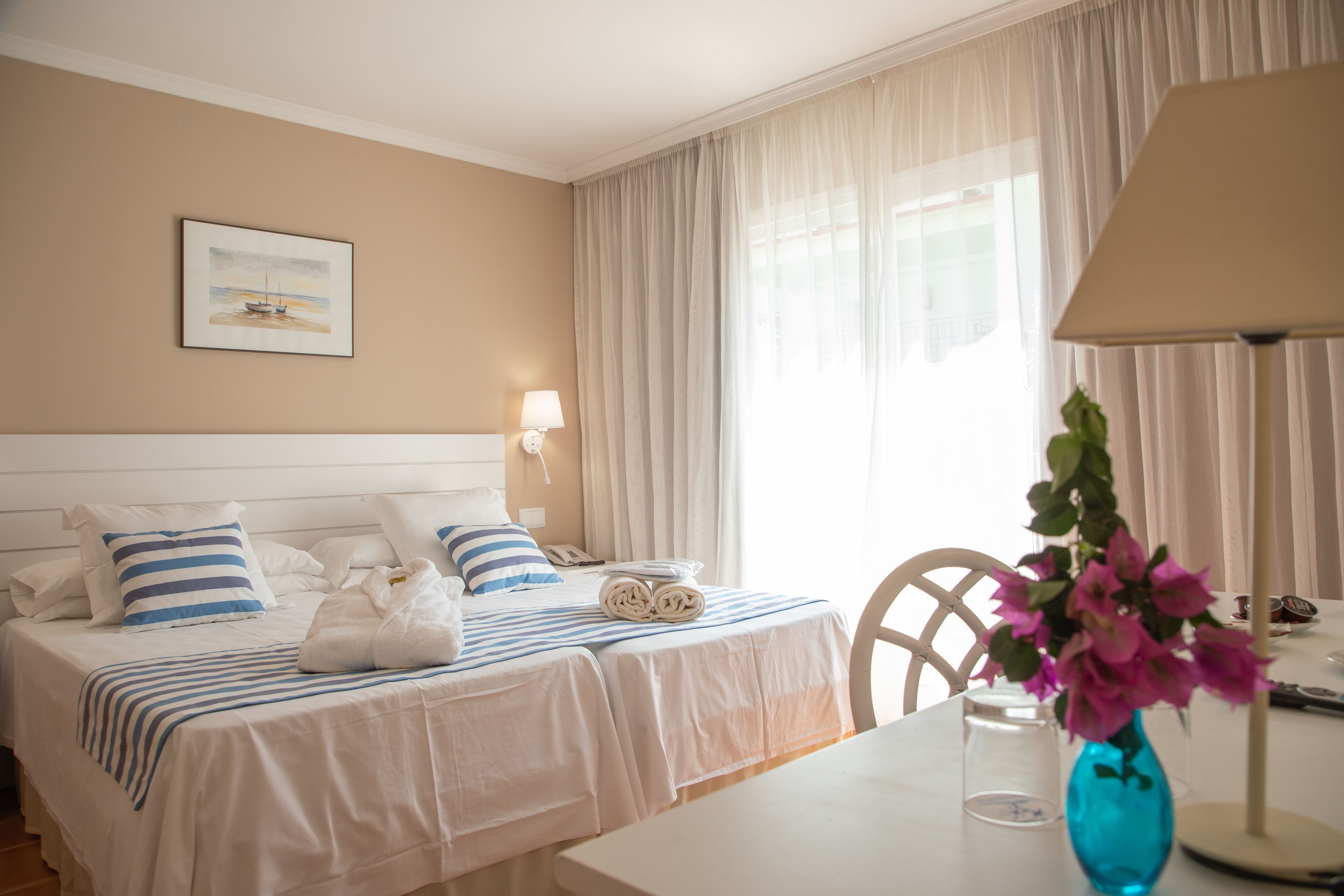 Ein Hotelzimmer mit Bett, Schreibtisch, Stuhl, Lampe, Blumen in einer Vase und einem Bild an der Wand.