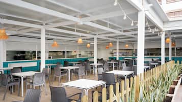An indoor dining area with tables and chairs, a kitchen in the background, and hanging pendant lights.