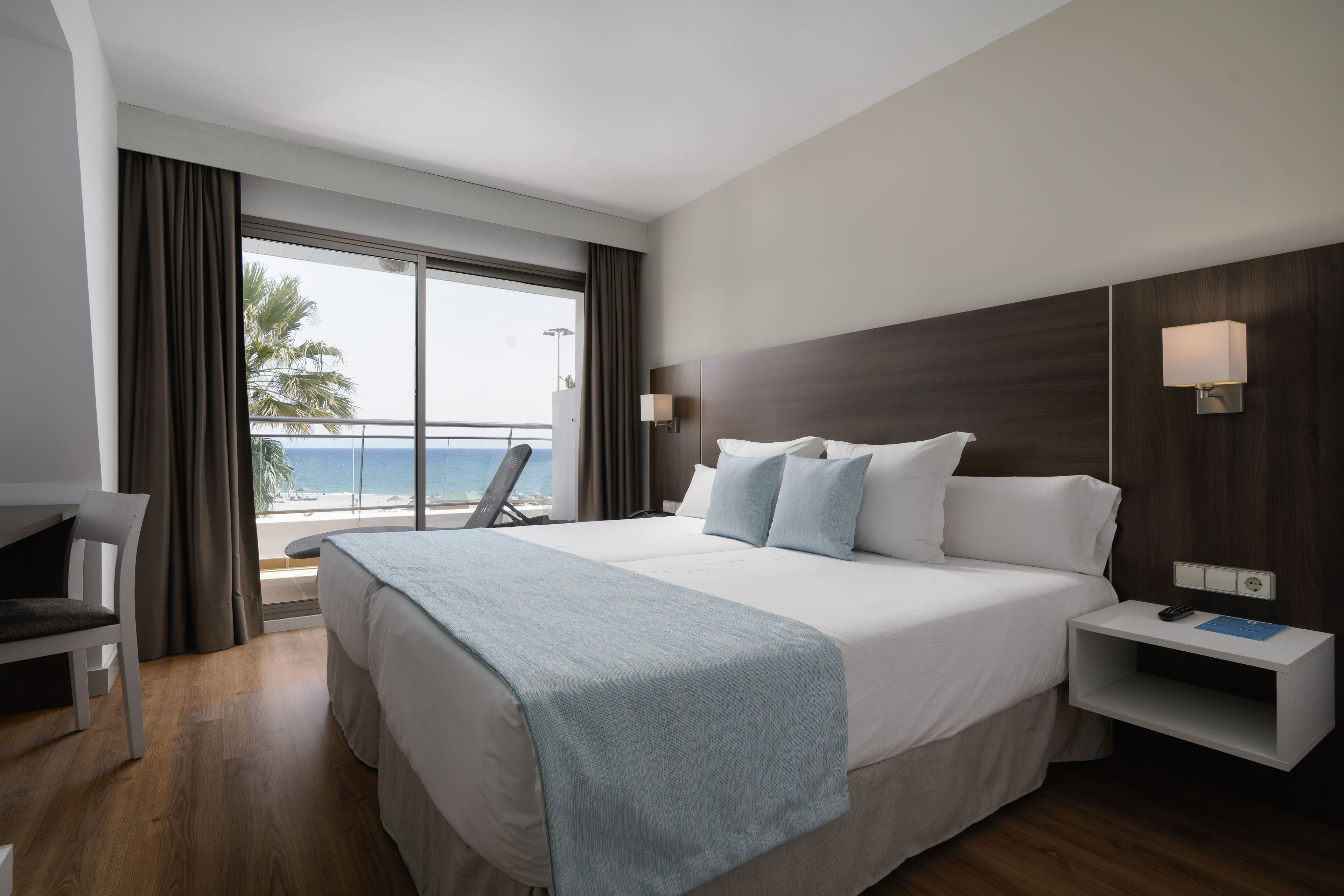 A hotel room with a large bed, a desk, a chair, a nightstand, and a view of the beach.
