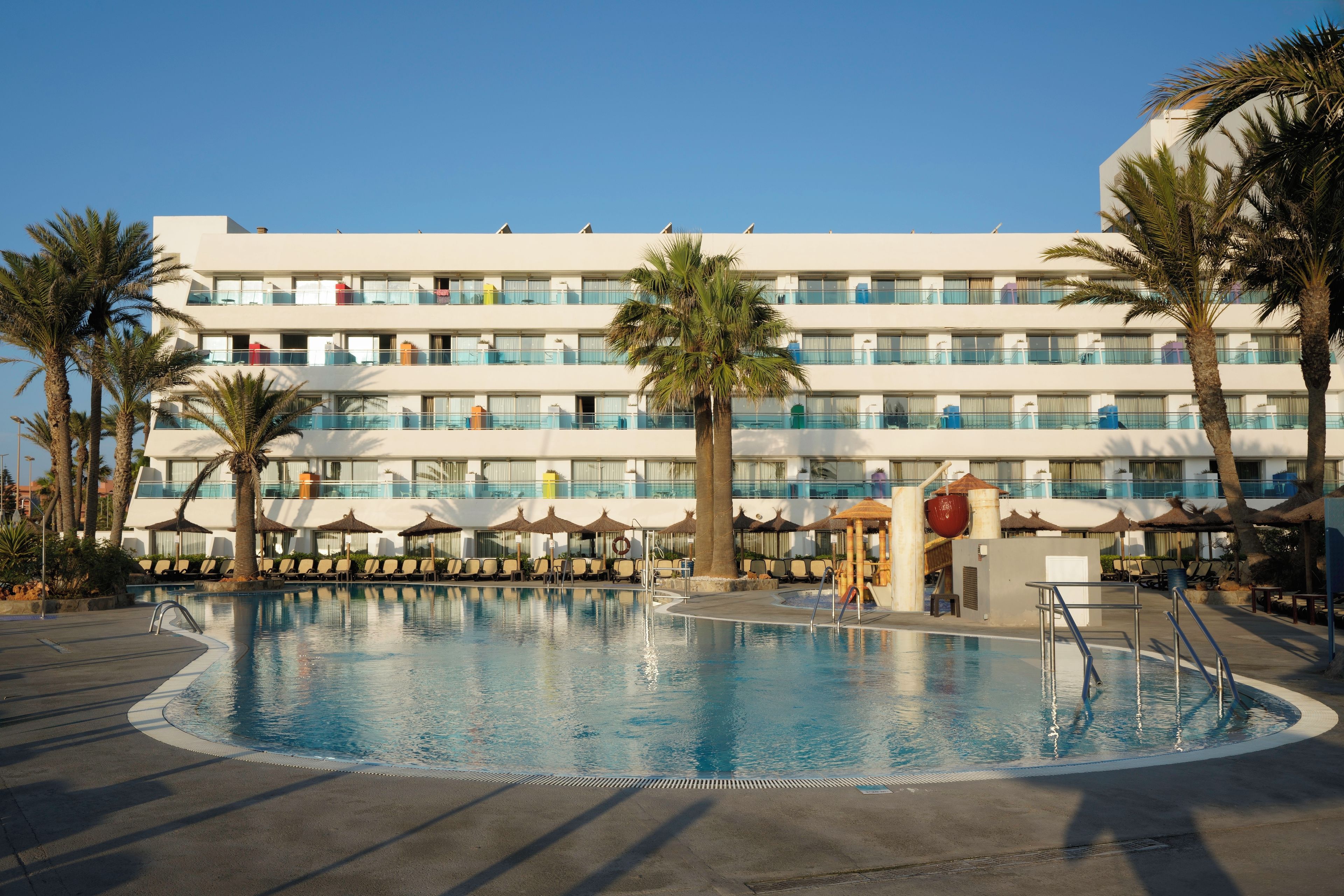 Un hotel con piscina, palmeras y un edificio de varios pisos con balcones.