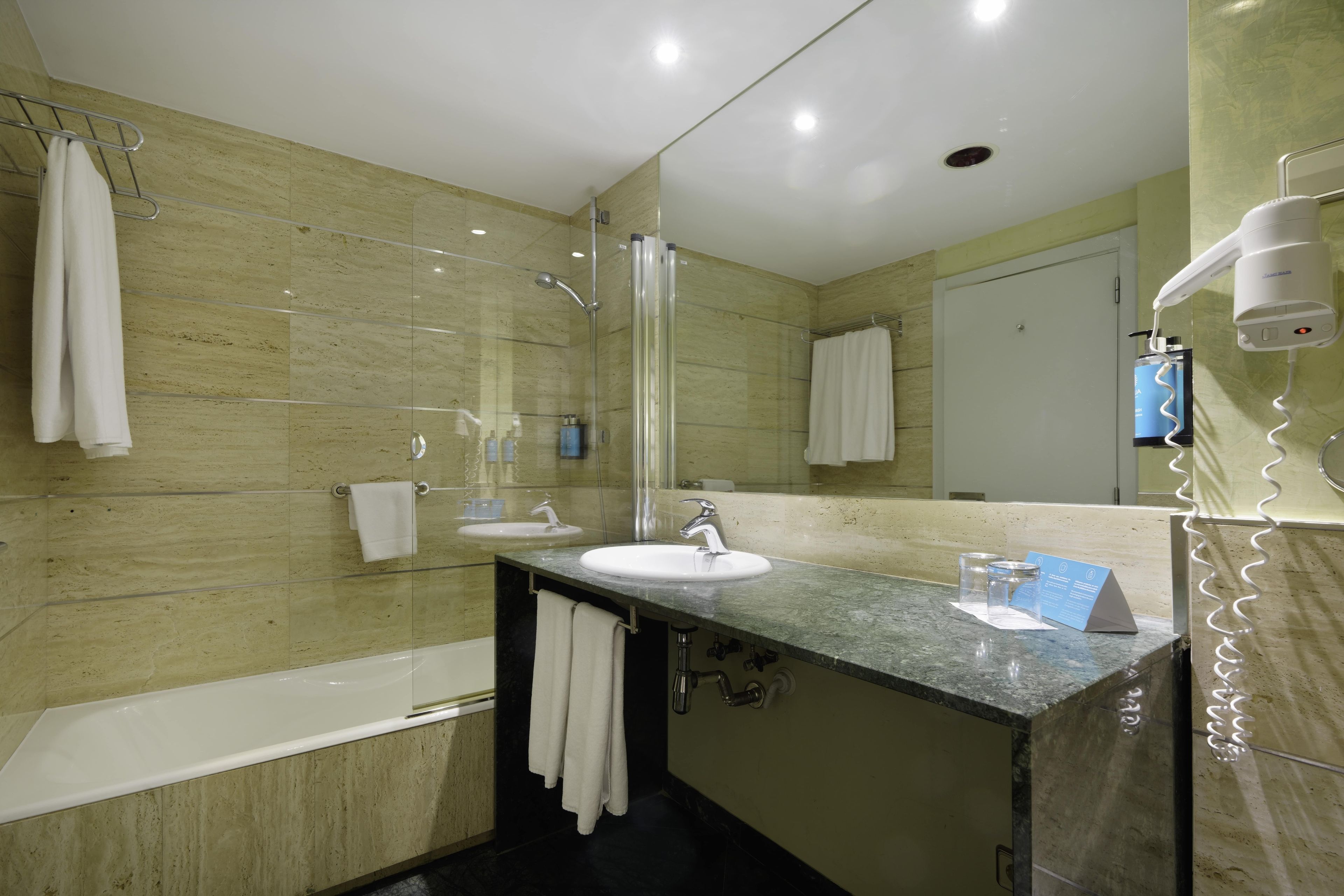 Un baño moderno con ducha envuelta en vidrio, un lavamanos con espejo y un secador de pelo sobre un soporte.