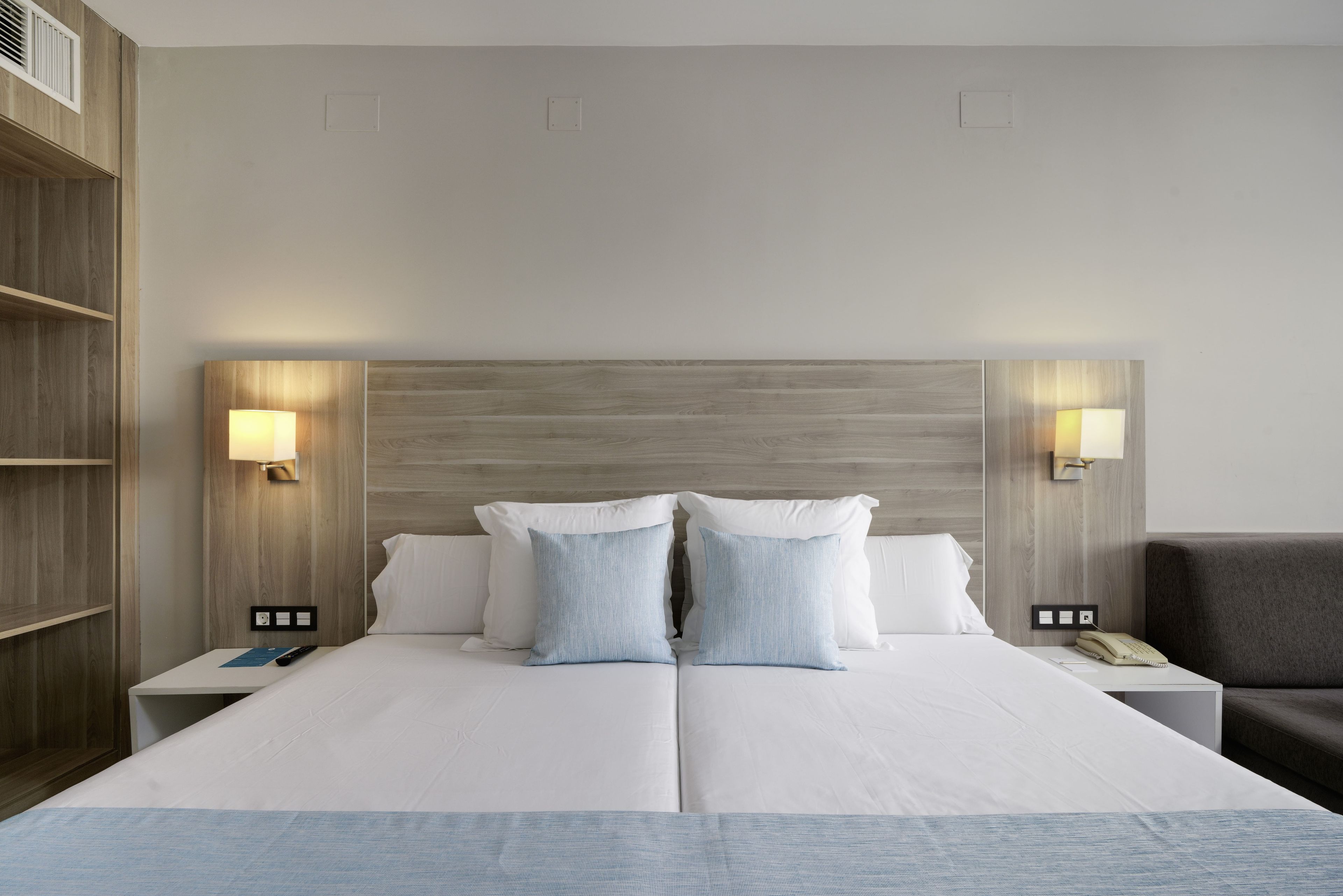 Una cama doble con dos almohadas, lámparas de noche, una mesita de noche con teléfono y un sillón gris.