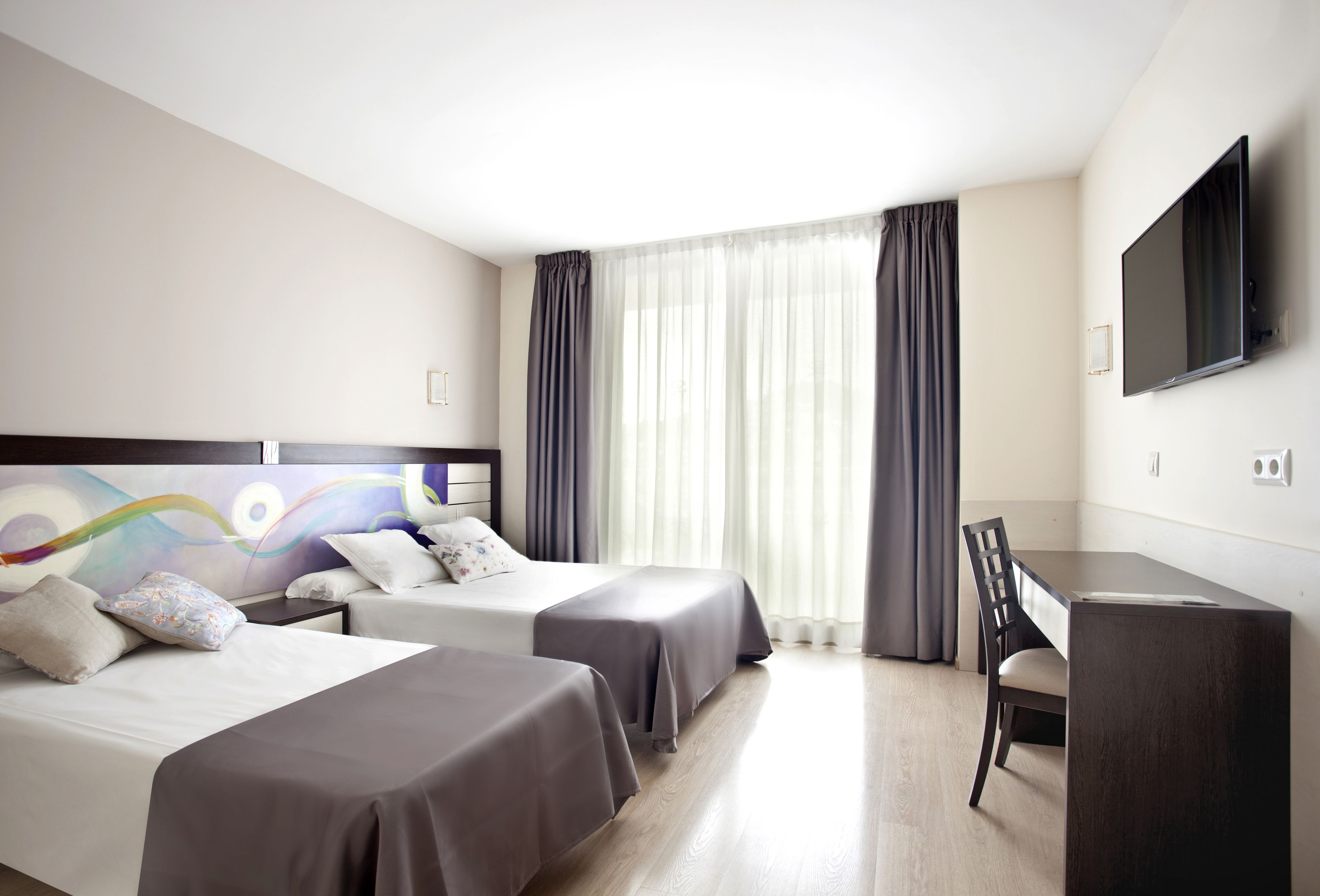 A hotel room with two beds, a desk, a chair, a TV, and a large window with curtains.