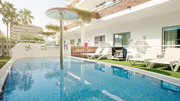 Una piscina con sillas de descanso y un toldo, rodeada por un edificio blanco con balcones.