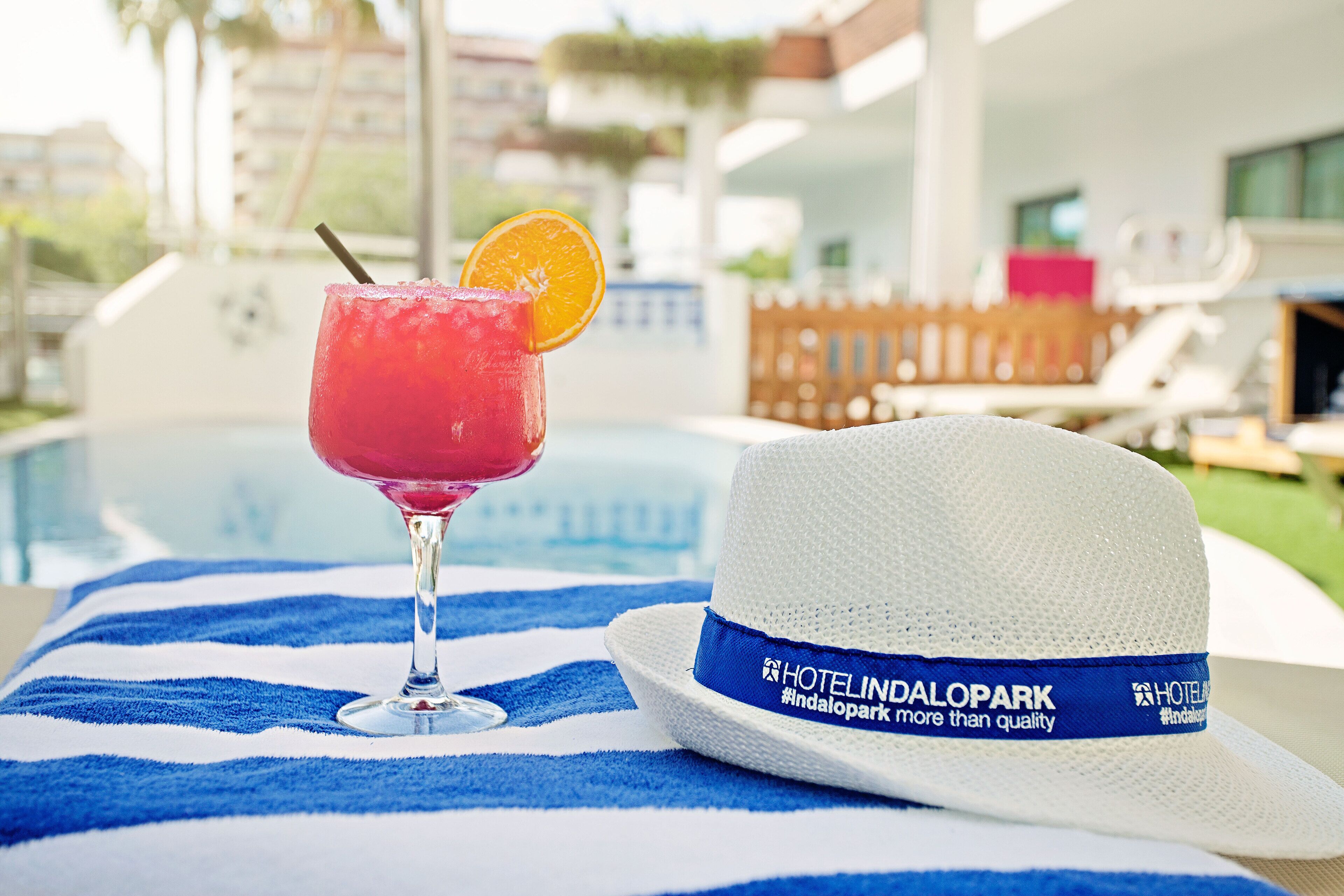 Un vaso de cóctel con una rodaja de naranja sobre una toalla azul y blanca, y un sombrero blanco con una cinta azul que dice "HOTELINDALOPARK #indalopark más que calidad".