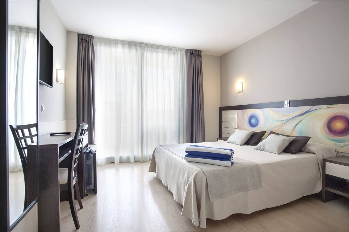 Habitación de hotel con cama, escritorio y silla. Hay un cuadro en la pared y una ventana con cortinas.