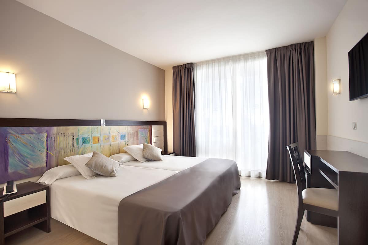 Habitación de hotel con una cama grande, un escritorio y una silla. Hay un cuadro en la pared, una lámpara y un televisor montado.