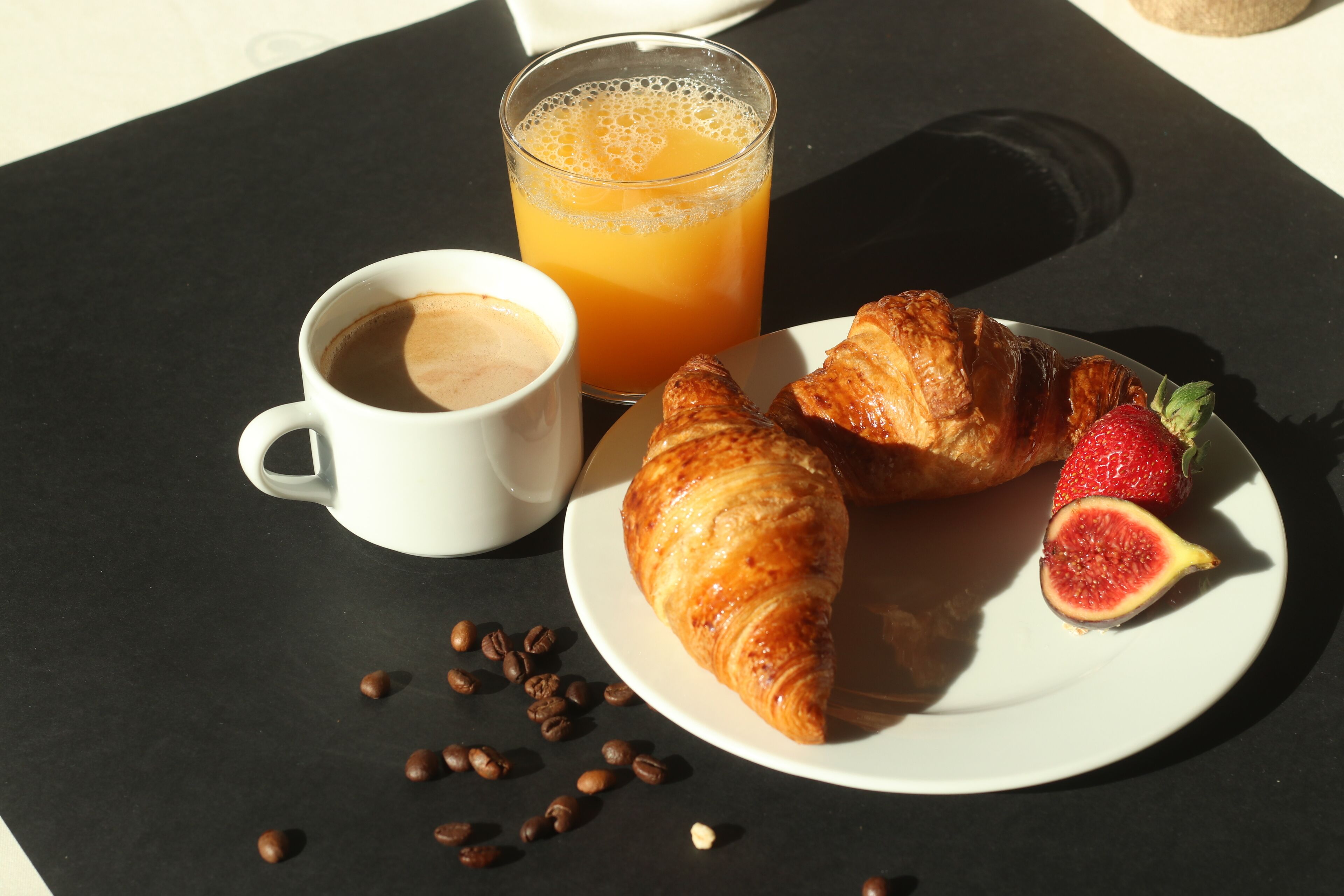 Un desayuno con un croissant, una fresa, un vaso de jugo de naranja y una taza de café sobre una mesa negra.