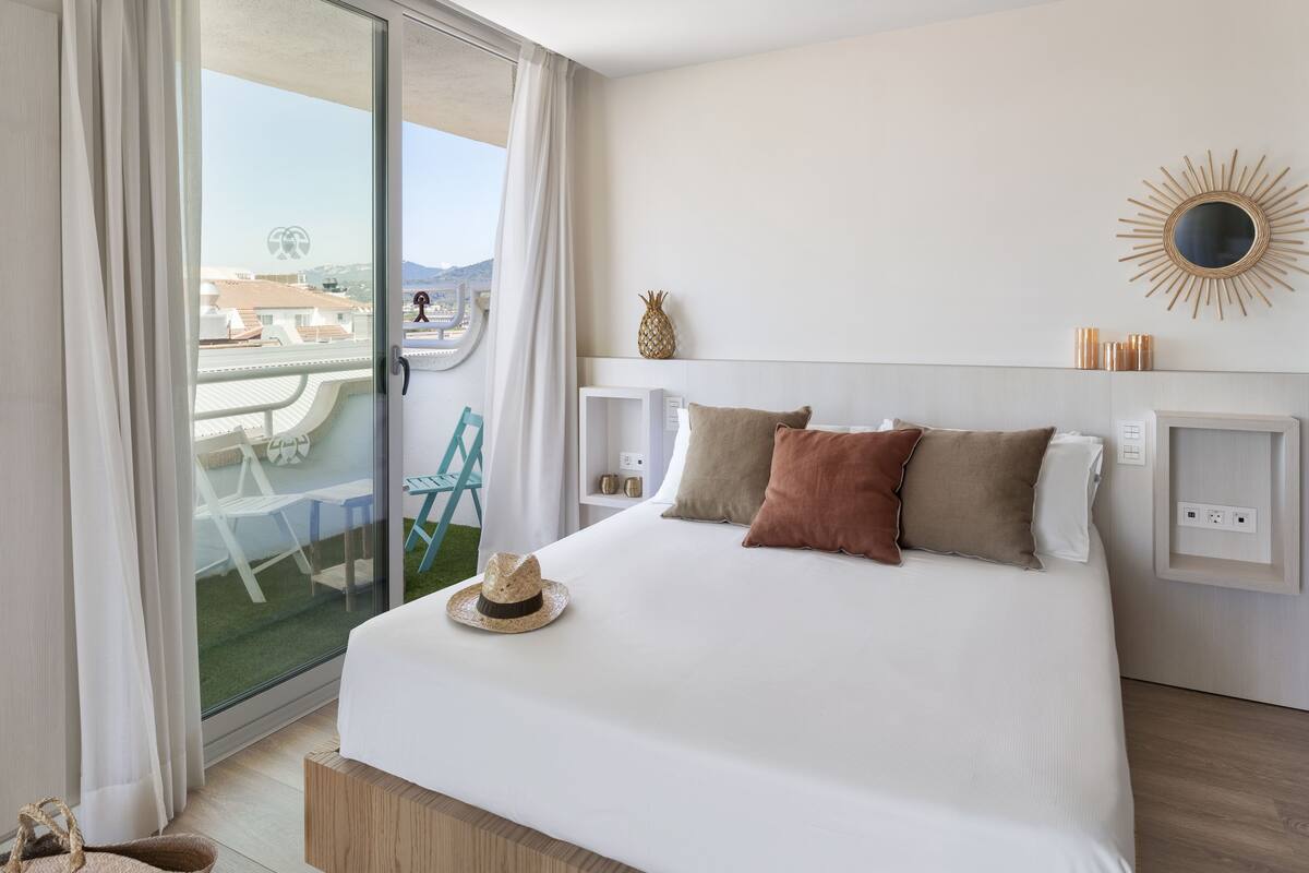 Habitación de hotel con una cama, un ventanal con cortinas, vista al exterior y un espejo con forma de sol en la pared.
