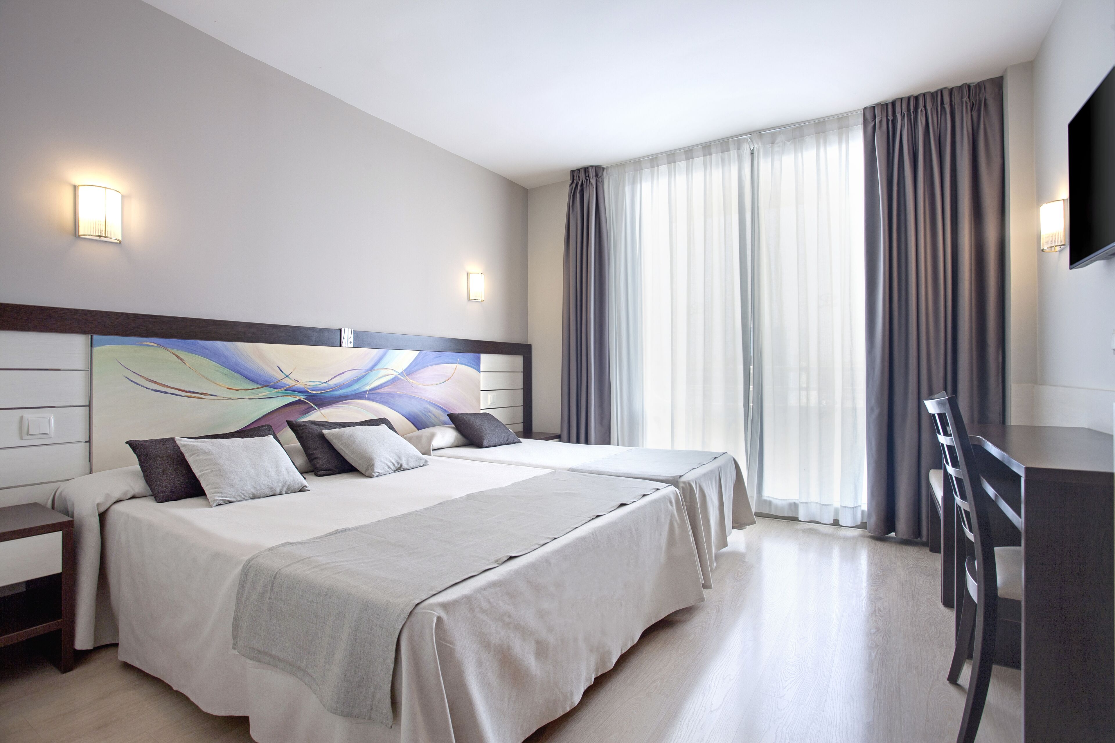 A hotel room with two beds, a desk, a TV, and a large window with curtains.