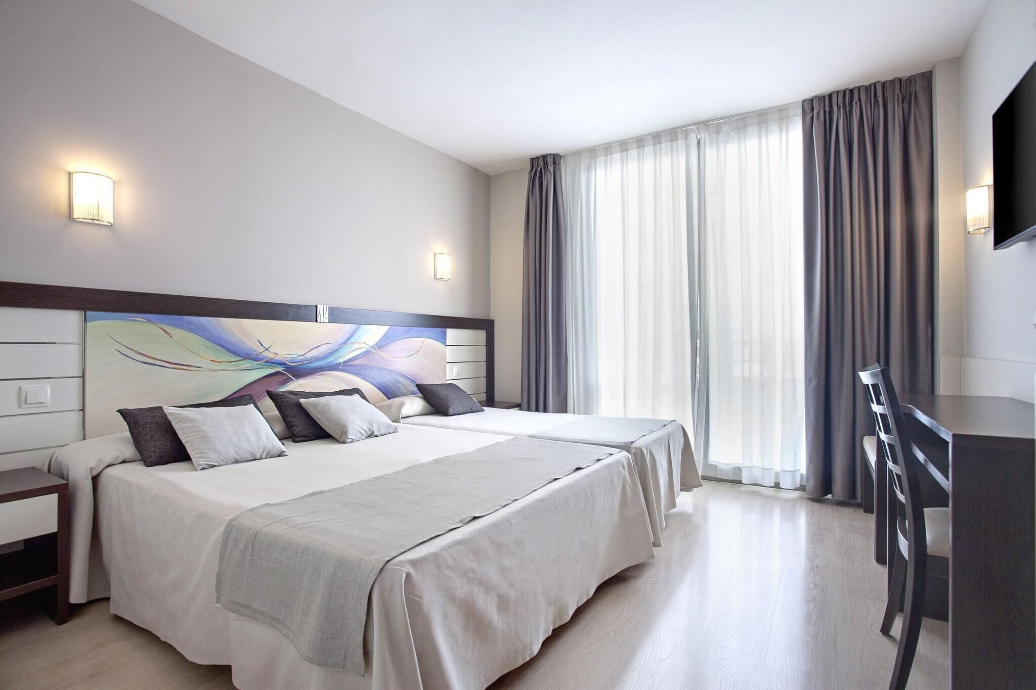 Una habitación de hotel con dos camas, un escritorio, un televisor de pantalla plana y un ventanal con cortinas.