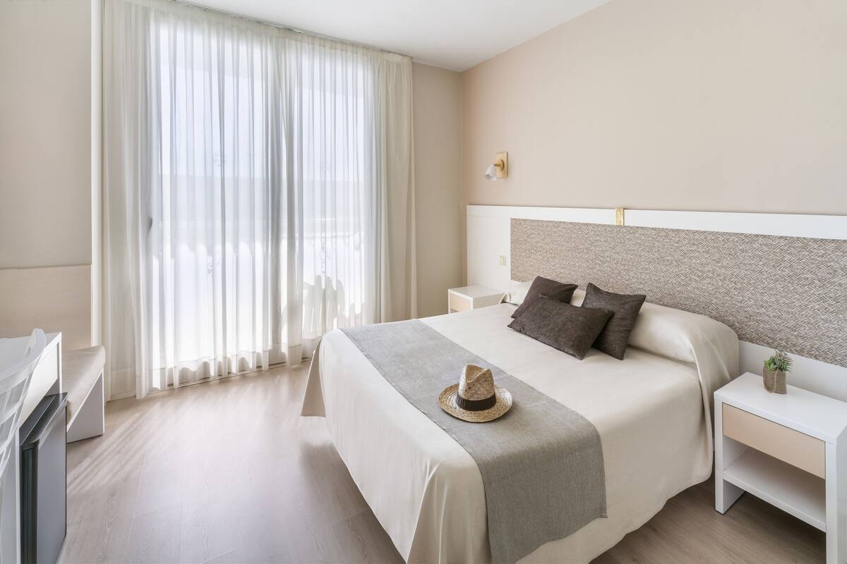 Una habitación de hotel impecable con una cama grande, una mesita de noche y un ventanal con cortinas traslúcidas.