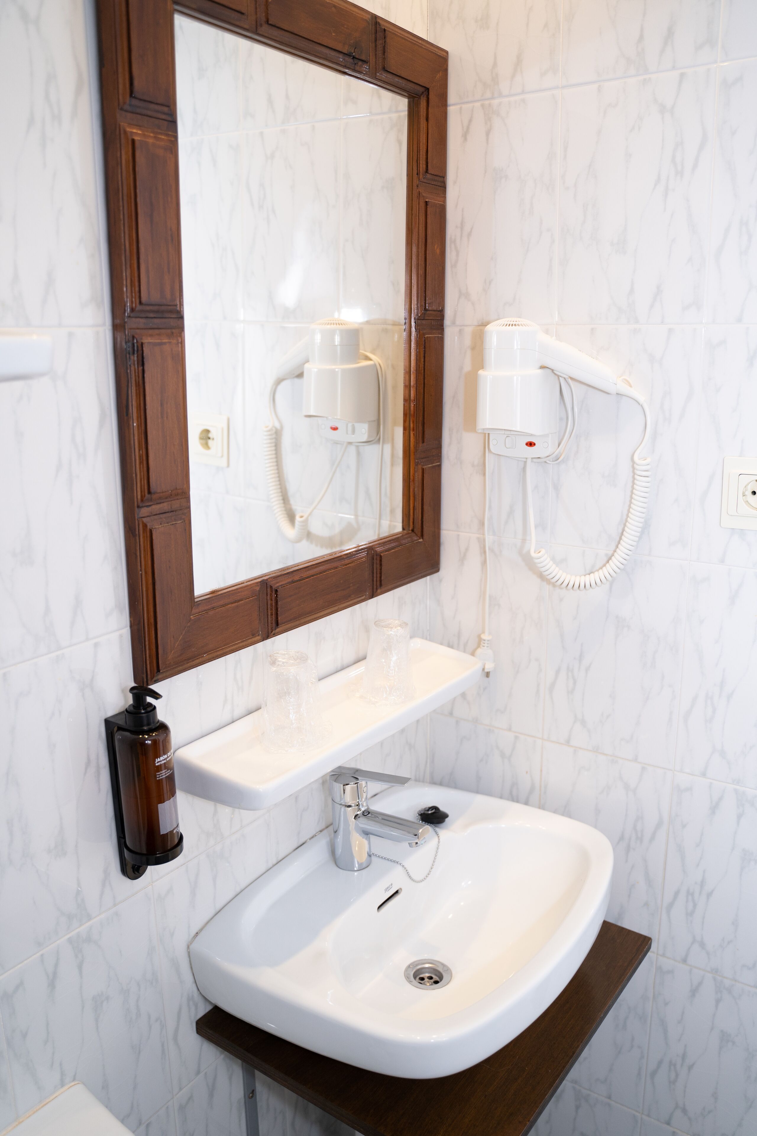 A bathroom with a marble wall, a wooden-framed mirror, a white sink, and a hairdryer mounted on the wall.