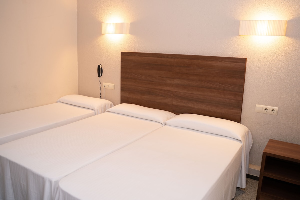 Une chambre d’hôtel avec deux lits simples, une tête de lit en bois, des lampes de chevet, un téléphone et une petite table de chevet en bois.