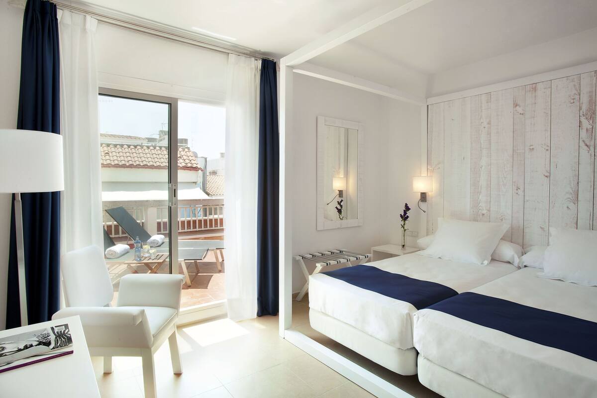 Une chambre d’hôtel avec deux lits, une chaise, une table et un balcon donnant sur des immeubles.