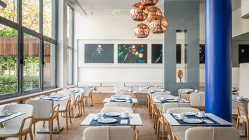 Un comedor moderno con mesas dispuestas para el servicio, ventanas amplias y obras de arte en las paredes.
