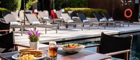 A dining table with a salad, sandwich, and drink, surrounded by lounge chairs and umbrellas by a pool.
