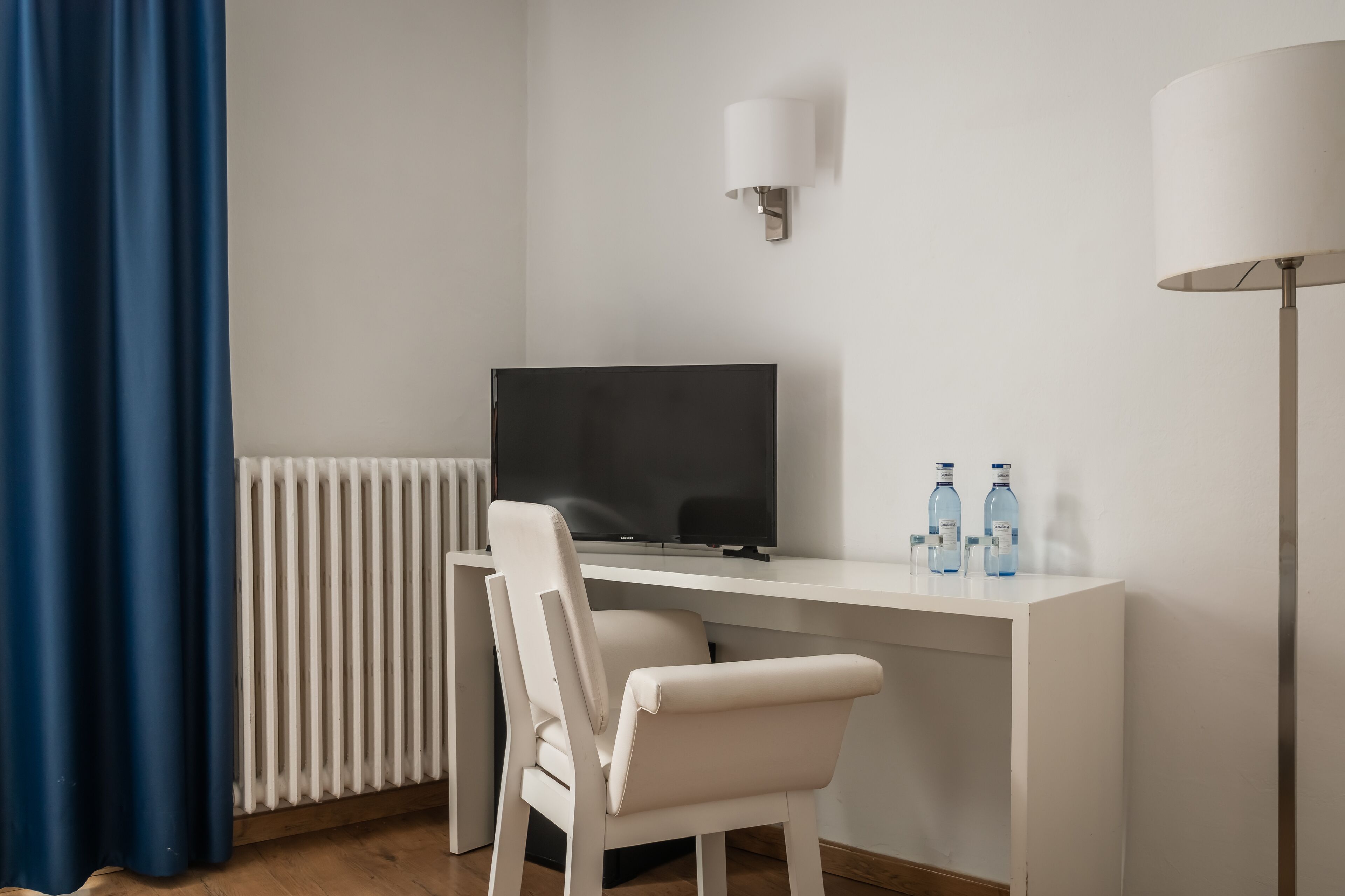 A hotel room with a white desk, a chair, a television, a radiator, a lamp, and two water bottles.