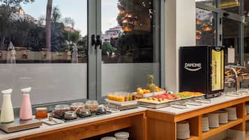 Un bufé de desayuno con diversas opciones de comida y bebidas, un refrigerador Danone y vistas al exterior a través de amplias ventanas.