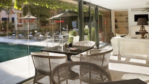 Un área de comedor exterior moderna con sillas de mimbre y una mesa de vidrio, con vistas a una piscina y un jardín.