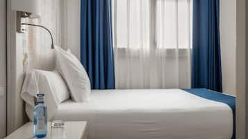 Una habitación de hotel con cama, una mesita de noche con una botella y un vaso, y cortinas azules.