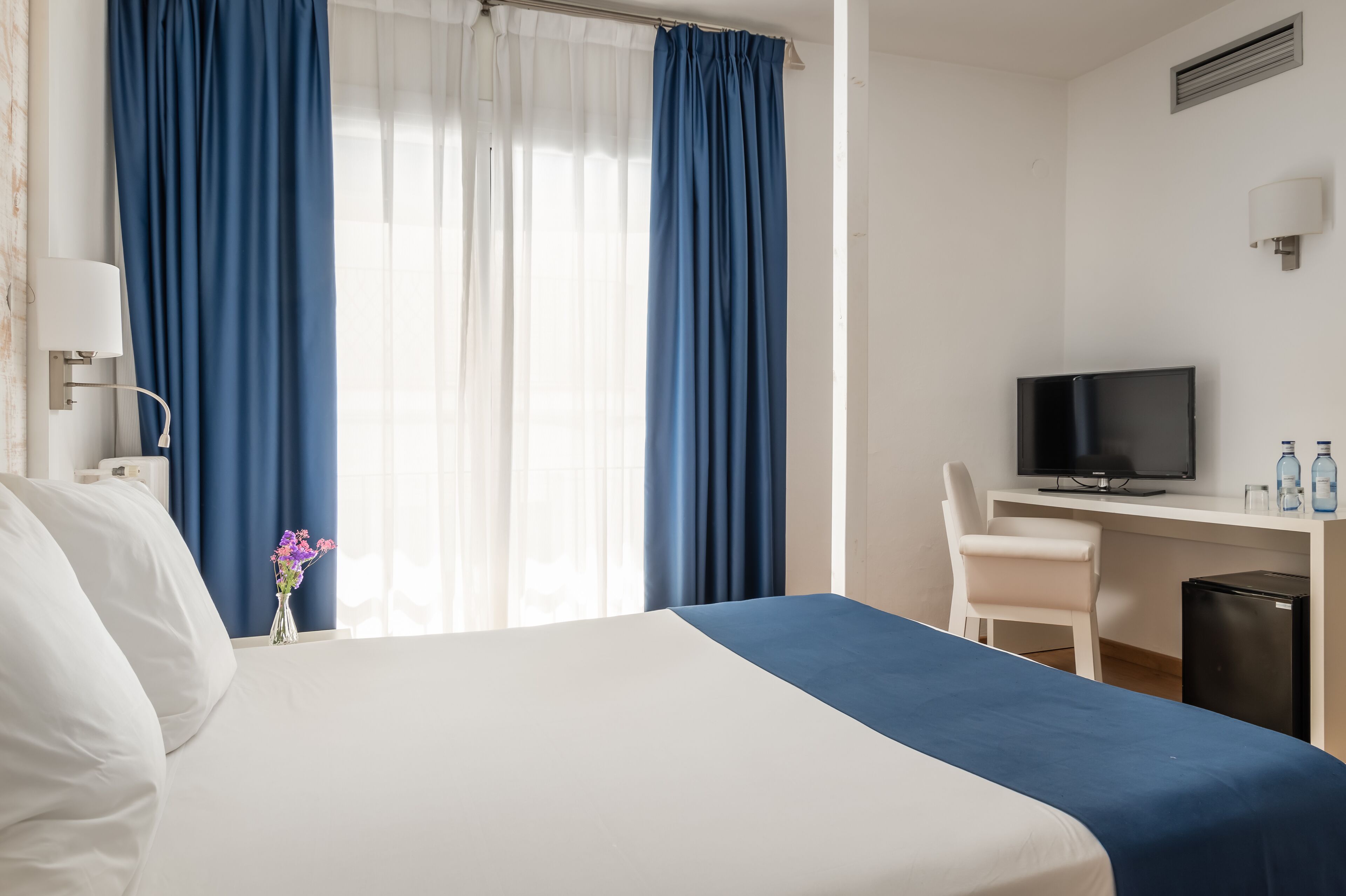Een hotelkamer met een bed, een bureau met een televisie, een stoel en een tafeltje met een bloevas.