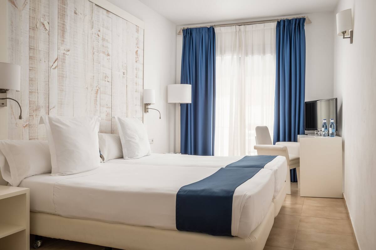 Une chambre d’hôtel avec un grand lit, un bureau et une chaise. Il y a une télévision sur le bureau et une fenêtre avec des rideaux.