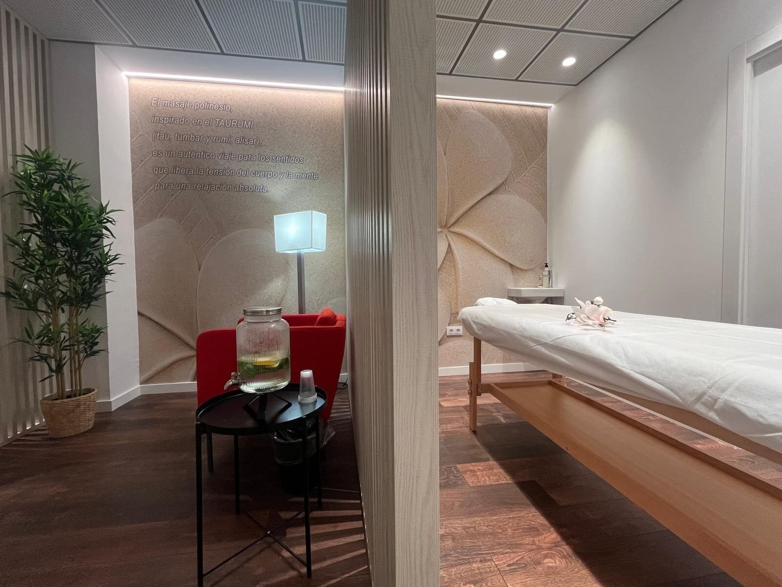 A modern room with a massage table, a small table with a lamp, and a wall with text about massage therapy.