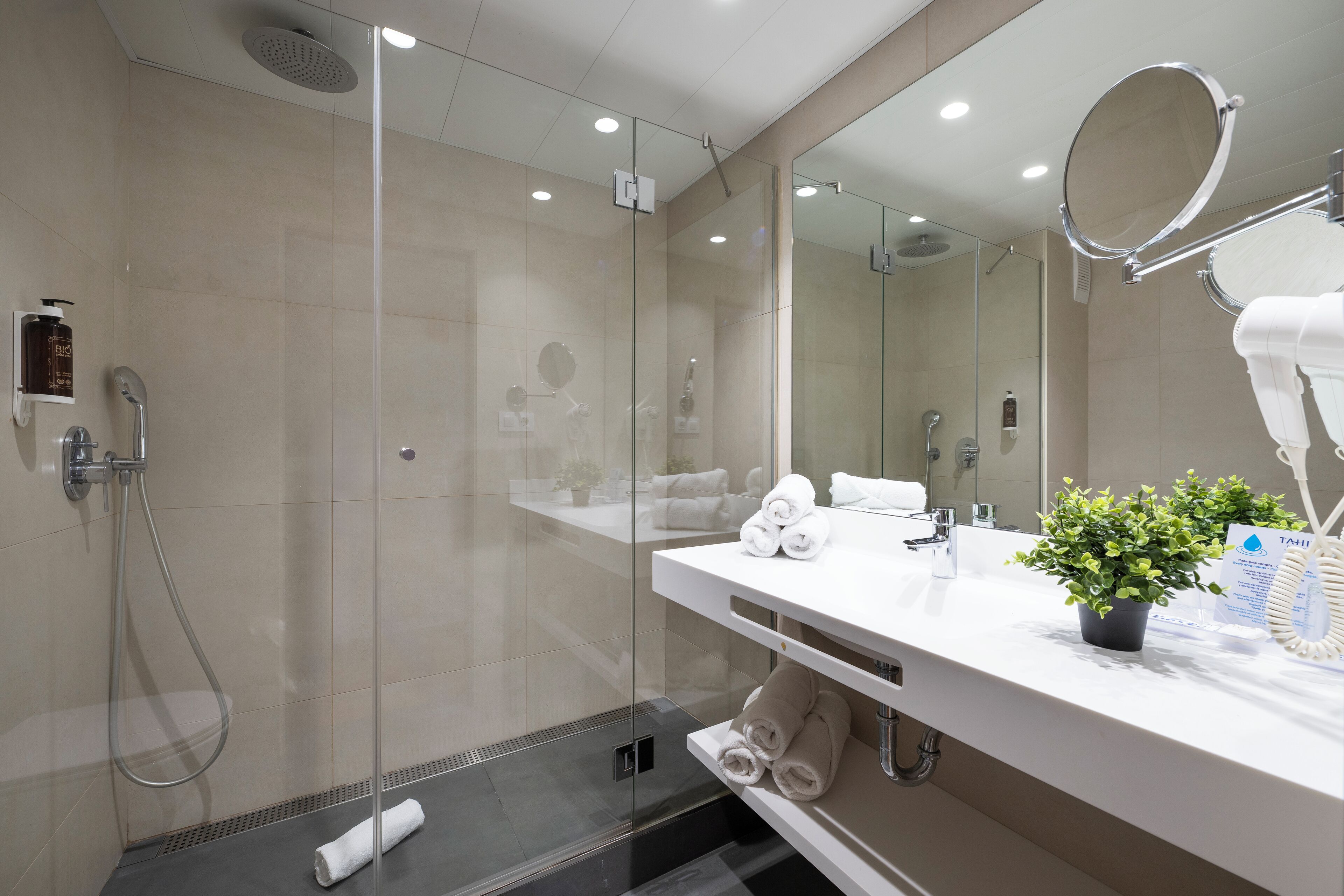 Un baño moderno con ducha envuelta en vidrio, un tocador blanco con espejo y una planta en maceta.